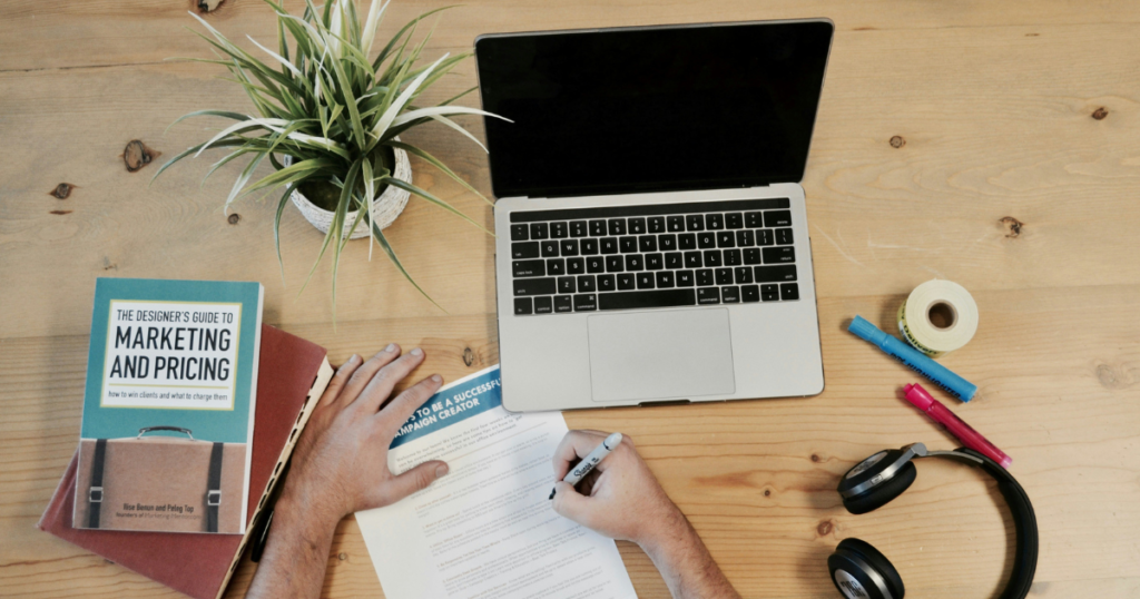 Laptop on a desk with an individual writing on paper.