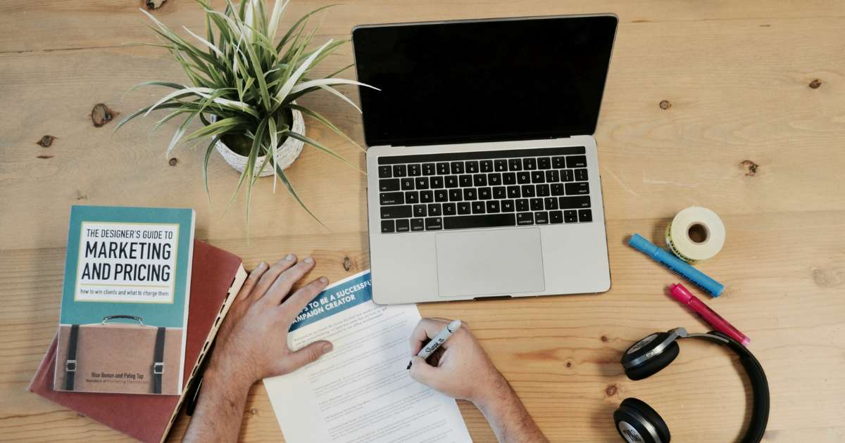 Laptop on a desk with an individual writing on paper.